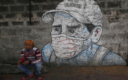 <p><strong>WAITING FOR ORDERS.</strong> A delivery rider sits beside a Covid-19-themed wall graffiti of a man wearing a face mask while waiting for orders from customers at Teacher's Village in Quezon City in this photo dated April 18, 2023. The Covid-19 positivity rate in the National Capital Region has reached 25.4 percent as of May 13, 2023. <em>(PNA photo by Joan Bondoc)</em></p>