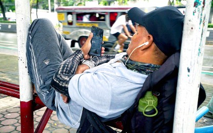 <p><strong>NOT A CARE. </strong>Unmindful of the movements around him, a man watches a movie on his mobile phone while comfortably lying under a waiting shed in Visayas Avenue, Quezon City on July 25, 2023. The public is warned of a new short message service scam that can evade automatic blocking tools by using unclickable links. <em>(PNA photo by Ben Briones)</em></p>