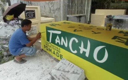 <p><strong>GRAVE HUMOR:</strong> Artist Jafryl Refran unveils his latest Tancho-pomade-inspired tomb painting for the Relleve family of Oas, Albay, who practices "grave branding" in this photo taken on Oct. 25, 2023. Every year, it has become a family tradition to paint their father's tomb with his favorite brands of shoes, liquor, food, and perfume, among other things. <em>(Photo courtesy of Jafryl Refran)</em></p>